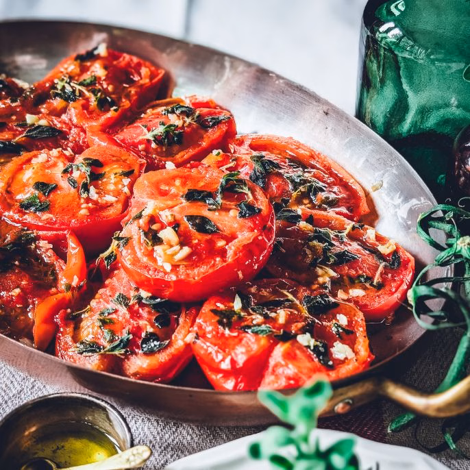 Pan of pan-fried tomatoes with fresh garden herbs on a table covered in a white tablecloth. Next to the pan some trinkets with salt and oil, fresh oregano and two empty places and cutlery